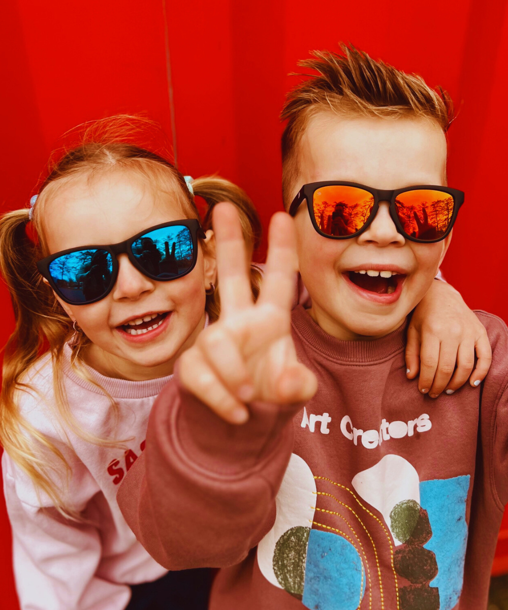 Twee lachende kinderen met kinderzonnebrillen. De ene kinderzonnebril heeft rode spiegelglazen en de ander heeft blauwe spiegelglaze.